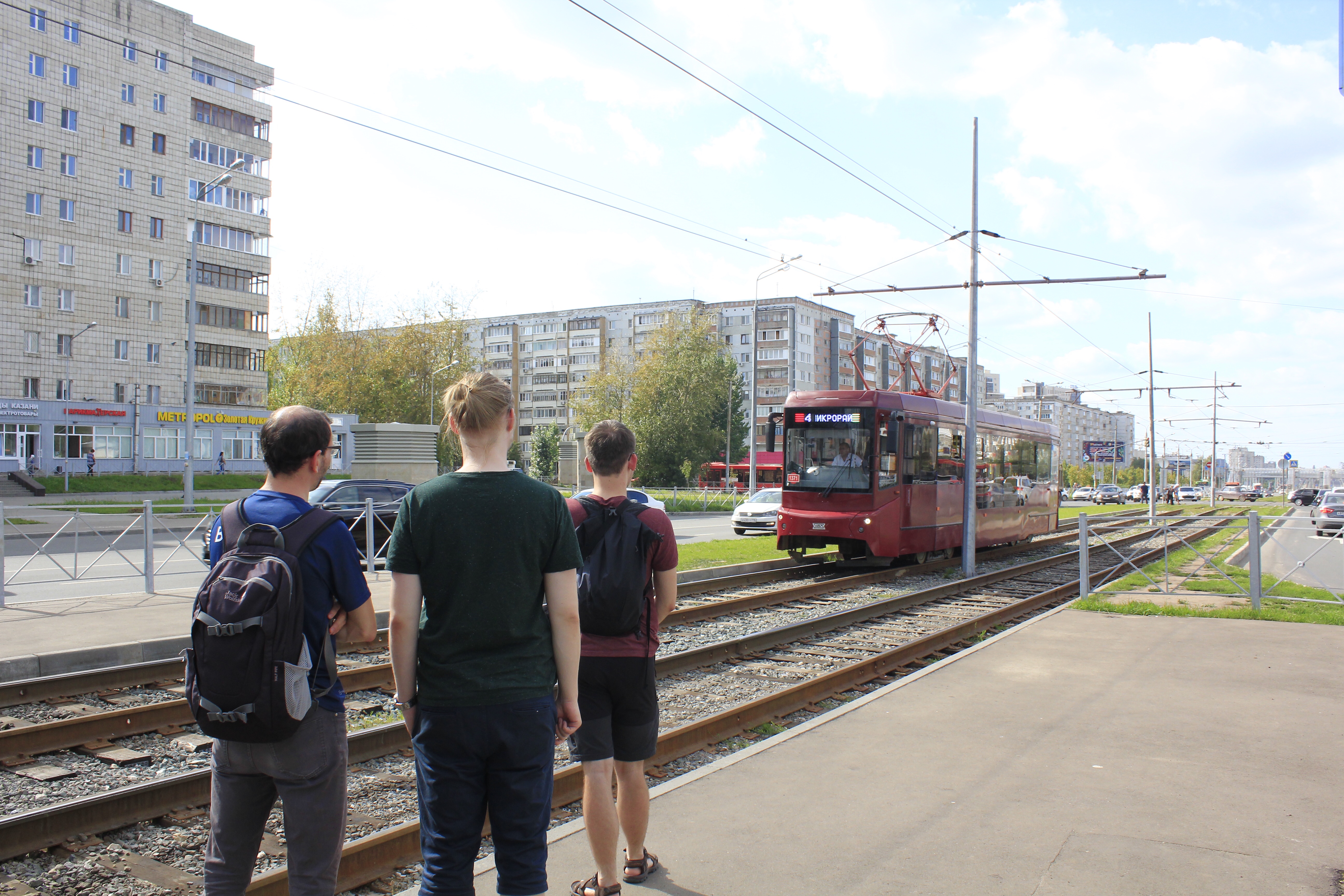 Torsten, Jan und Julius stehen an einer  Straßenbahnhaltestelle und gucken einer einfahrenden Straßenbahn zu. Im Hintergrund stehen viele Wohnblöcke 